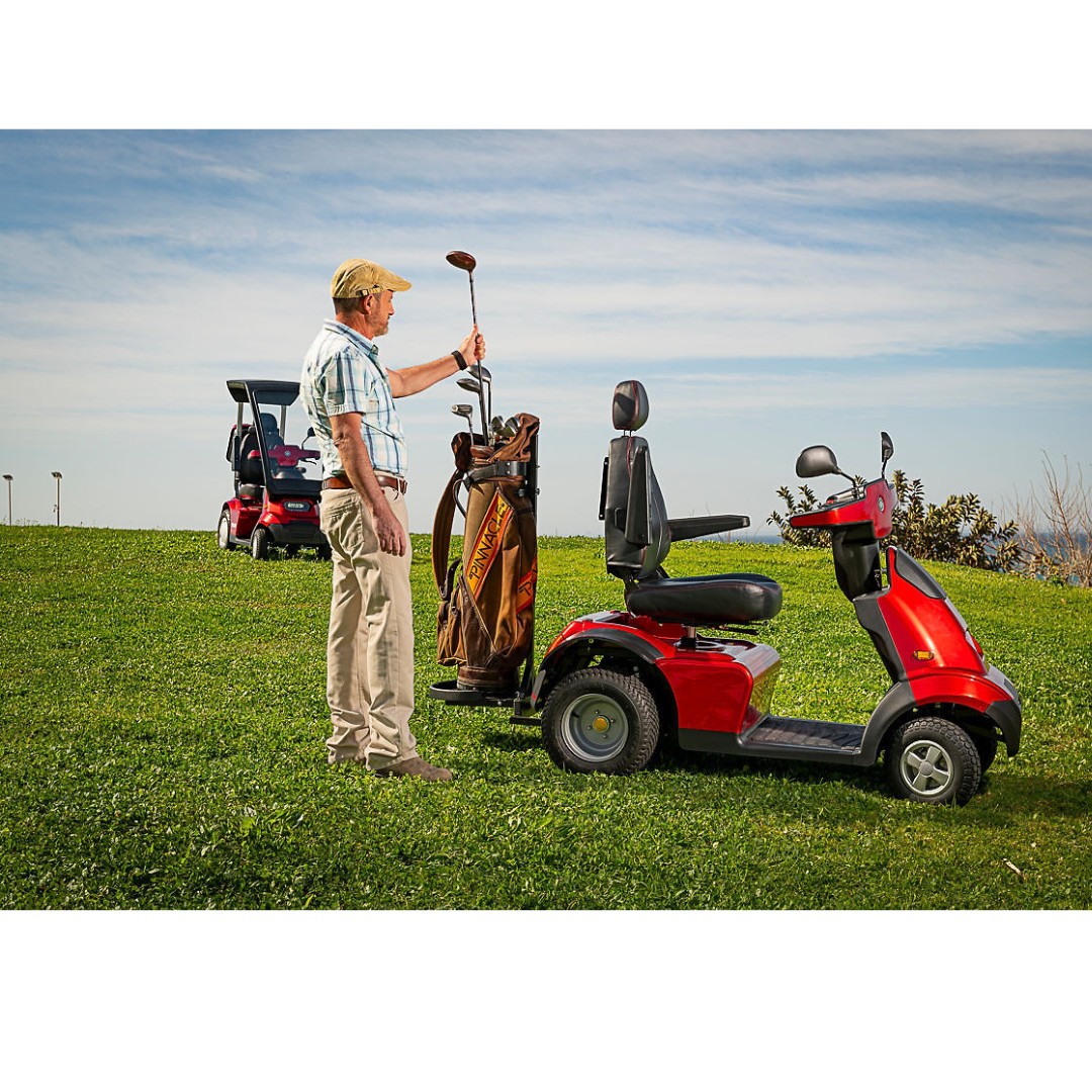 An older man holds a golf club on a grassy hill next to a red Afiscooter featuring Afikim Electric Vehicle Accessories with a custom golf bag. Another red scooter is in the background under a blue sky sprinkled with clouds.
